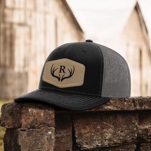 May include: Black and gray trucker hat with a tan leather patch featuring a stylized antler design and the letter "R". The hat has a curved brim and a mesh back. The hat is sitting on a brick surface.