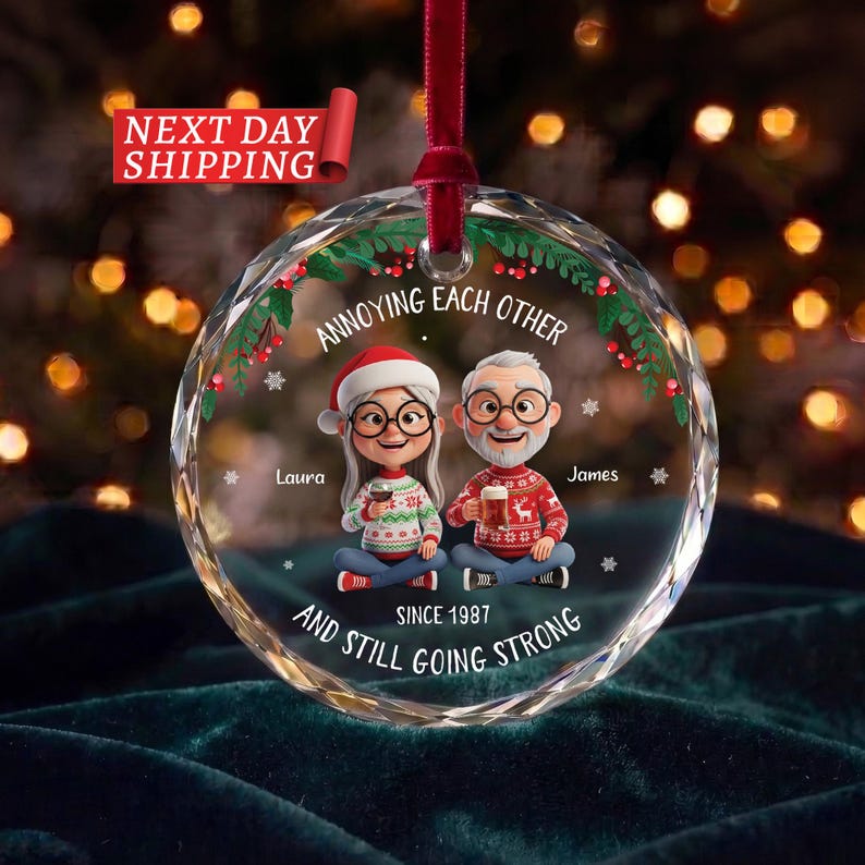 May include: A round, clear glass Christmas ornament featuring a cartoon illustration of a couple wearing holiday jumpers and glasses. The text reads: ANNOYING EACH OTHER, Laura, James, SINCE 1987, AND STILL GOING STRONG. It has a red ribbon hanger.