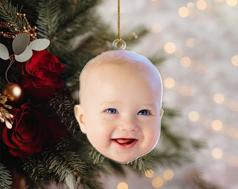 Puede incluir: Adorno navideño con la cara sonriente de un bebé con ojos azules, suspendido por un hilo dorado. El fondo muestra un árbol de Navidad con rosas rojas y adornos dorados. Una decoración festiva para las fiestas.