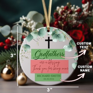 May include: A 3-inch round white ceramic ornament with a green and white floral design. The ornament features a green, white, and red banner with the text "Godfathers are a blessing. Thank you for being mine."  The ornament also includes a green banner with the text "RICHARD SMITH 06.18.2024".