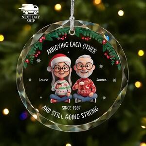 May include: A round, clear, faceted glass Christmas ornament featuring a cartoon illustration of an older couple in holiday sweaters. The text reads: ANNOYING EACH OTHER, Laura, James, SINCE 1987, AND STILL GOING STRONG. It includes a silver hanging cord.