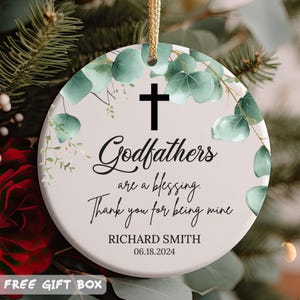 May include: A white round Christmas ornament with a green floral design and a black cross. The ornament reads "Godfathers are a blessing. Thank you for being mine" and "RICHARD SMITH 06.18.2024".