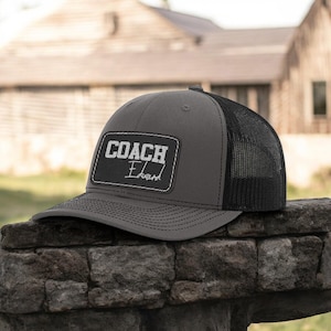 May include: A gray and black trucker hat with a black mesh back. The hat has a white patch that says "COACH Edward" on it.