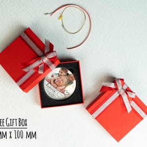 May include: A round white ceramic ornament featuring a printed photograph of two people, with the text "heaven" and "in loving memory" inside a 100 mm x 100 mm red gift box with a silver glitter ribbon, accompanied by a thin gold hanging cord.