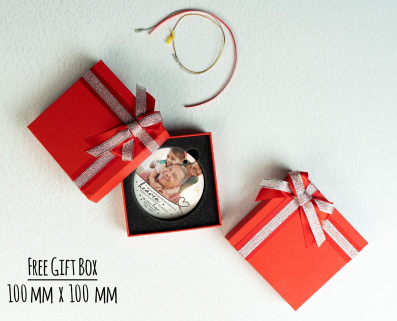 May include: A round white ceramic ornament featuring a printed photograph of two people, with the text "heaven" and "in loving memory" inside a 100 mm x 100 mm red gift box with a silver glitter ribbon, accompanied by a thin gold hanging cord.