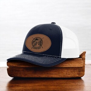 May include: A navy blue and white trucker hat with a brown leather patch featuring a fish and crossed fishing rods. The patch also includes the name "EDWARD".