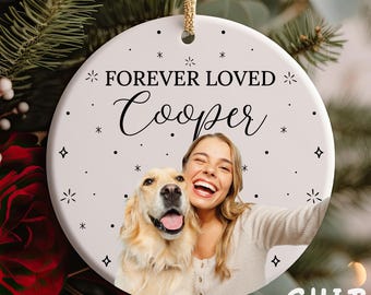 May include: A round white ceramic ornament featuring a gold hanging string. The text FOREVER LOVED and Cooper is printed in black. Below the text is a color photograph of a person smiling next to a golden retriever dog. Small black star icons decorate the surface.