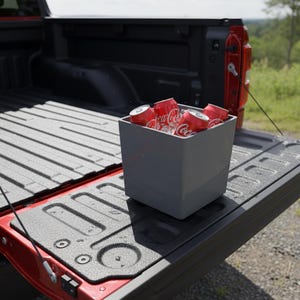 May include: A gray cooler filled with red Coca-Cola cans and ice sits in the bed of a red pickup truck. The truck bed is black with a textured surface. The scene is outdoors with green foliage in the background.