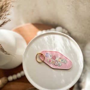 Peut inclure: Porte-clés rose en forme de clé de motel avec le texte "Bookish Babe" en lettres colorées.