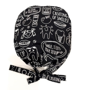 May include: Black and white scrub hat with a dental theme. The hat has a tie on the back and features illustrations of teeth, toothbrushes, apples, and the text "Be proud of your smile", "Creating of smiles!" and "Smile, it lets your teeth breathe".