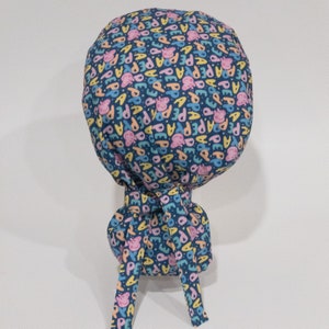 May include: A blue surgical scrub hat with a colorful pattern of pink pigs and the word "Peppa" repeated. The hat has a tie that can be tied at the back of the neck.