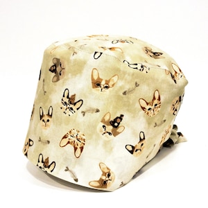 以下が含まれることがあります： 茶色の猫と魚の骨の模様がついたベージュのスクラブハット。帽子は後ろにリボンが付いています。