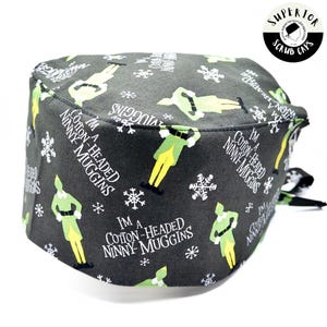 May include: A dark grey scrub cap featuring a repeating pattern of green and yellow elf figures, snowflakes, and the text "I'M A COTTON-HEADED NINNY MUGGINS" in white. The cap has a black tie.