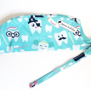 May include: A turquoise blue surgical scrub cap with a white and pink tooth pattern. The cap has a tie and a label that says "Superior Scrub Caps".