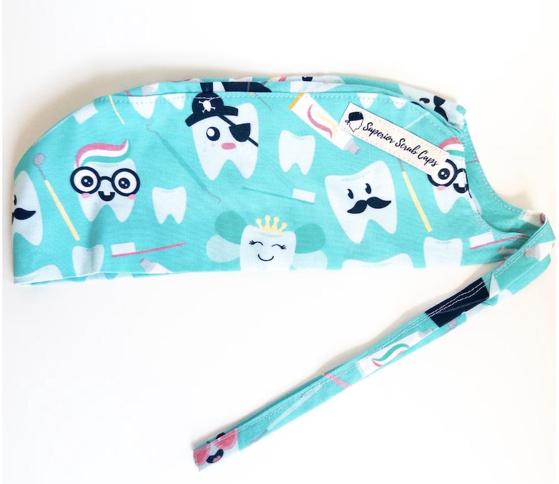 May include: A turquoise blue surgical scrub cap with a white and pink tooth pattern. The cap has a tie and a label that says "Superior Scrub Caps".