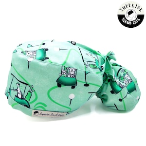 May include: Green scrub cap with a golf theme. The cap features a white cartoon dog riding in a golf cart with a golf club. The cap has a tie on the back and the text "Superior Scrub Caps" is printed on the bottom.