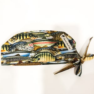May include: A patterned surgical scrub cap featuring a variety of fish illustrations in shades of brown, yellow, blue, and green. The cap has a tie in the back and a small tag that reads "Superior Scrub Caps".
