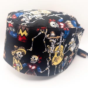 May include: Black scrub hat featuring a colourful pattern of skeletons playing musical instruments. The design includes a skeleton playing a guitar, a skeleton playing a drum, and a skeleton playing a trumpet. The hat is made from a soft, comfortable fabric.
