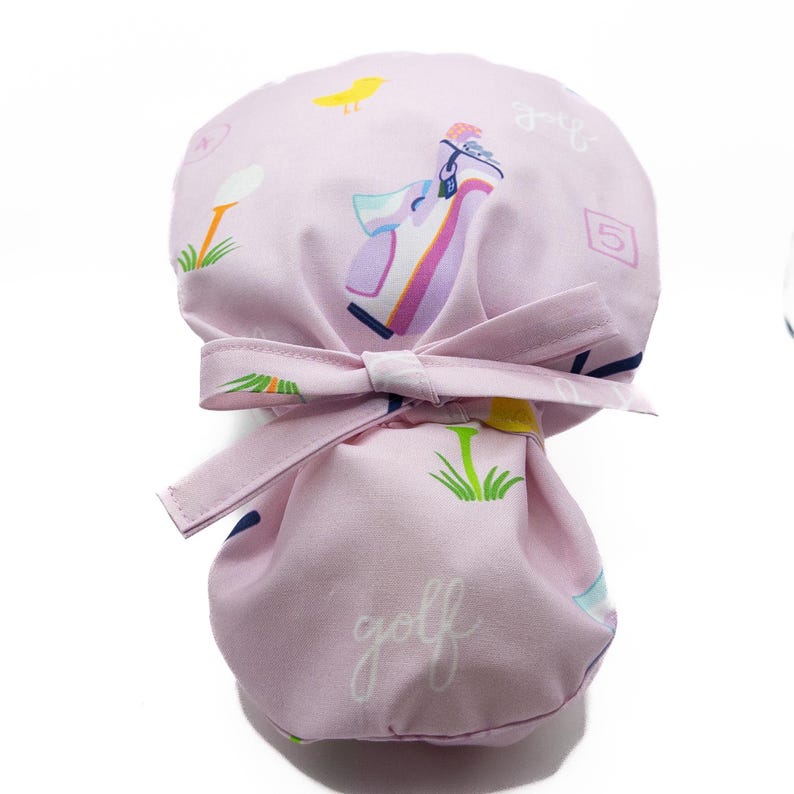 Pu&ograve; includere: Un cappello da chirurgo rosa chiaro con un motivo a tema golf. Il cappello presenta illustrazioni di mazze da golf, palline da golf, uccelli e la parola "golf" in corsivo. Ha una chiusura a fiocco sul retro.