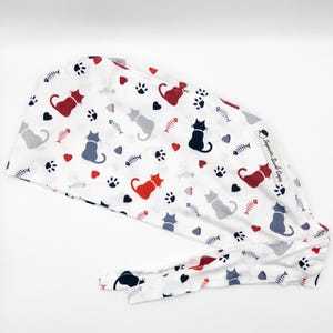 Op de afbeelding: Een witte chirurgische scrubcap met een rode, grijze en zwarte kattenprint. De cap heeft een strik aan de achterkant en een klein label met de tekst "Surgical Scrub Caps".