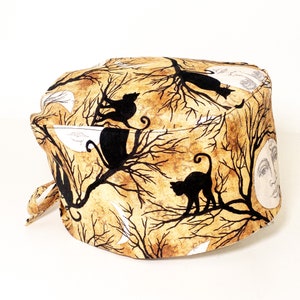 May include: A tan and brown scrub hat with a black cat and moon design. The hat has a tie on the side.