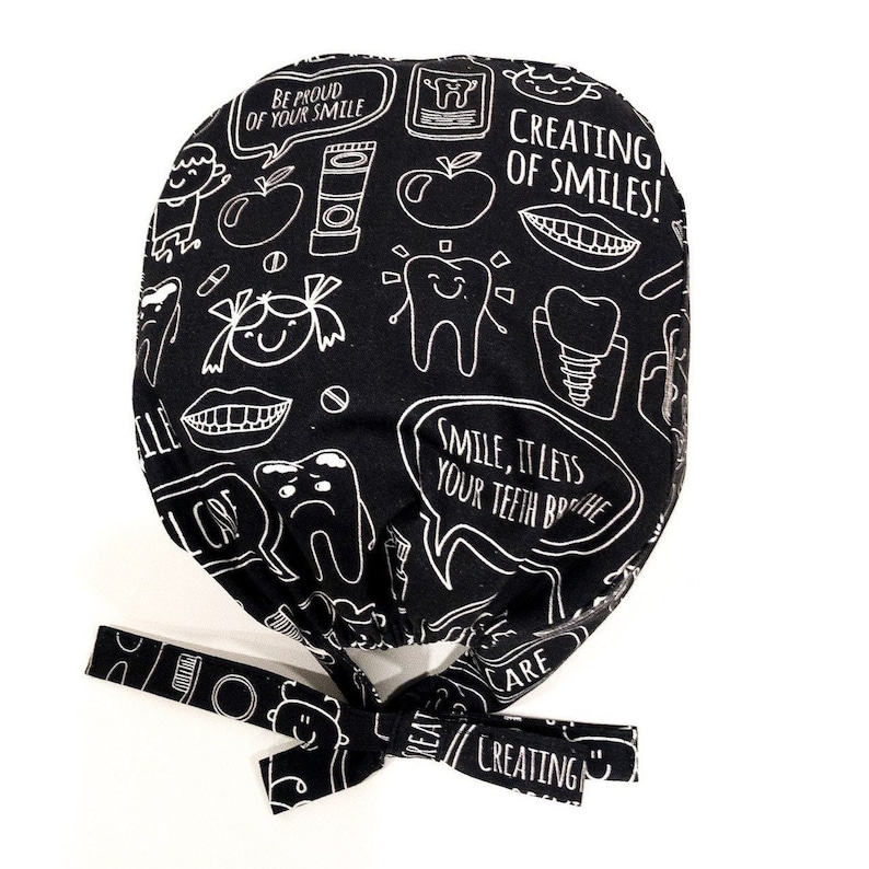 May include: Black and white scrub hat with a dental theme. The hat has a tie on the back and features illustrations of teeth, toothbrushes, apples, and the text "Be proud of your smile", "Creating of smiles!" and "Smile, it lets your teeth breathe".