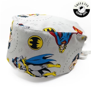 Puede incluir: Un gorro de quirófano gris con un estampado de Wonder Woman, Superwoman, Batwoman y un logotipo de Batman. El gorro tiene un lazo en la parte posterior. El texto "SUPERIOR SCRUB CAPS" está impreso en un círculo blanco en la esquina superior derecha.