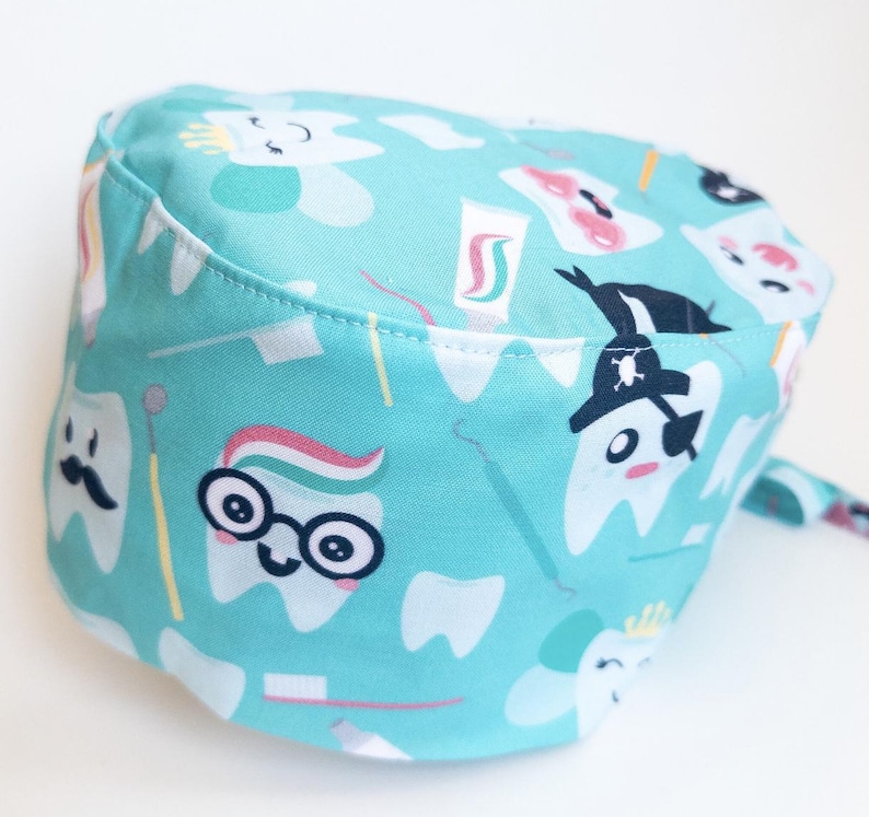 May include: A turquoise blue scrub hat with a fun pattern of cartoon teeth, toothbrushes, and toothpaste. The teeth have different expressions, including a pirate tooth with an eye patch.