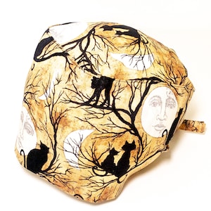 May include: A scrub cap with a Halloween design. The fabric features a repeating pattern of black cats, moons, and bare tree branches on a tan background. The cap has a tie at the back.
