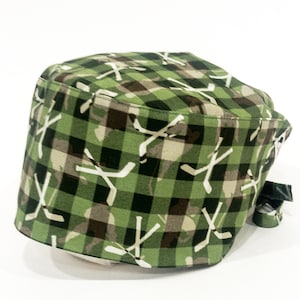 May include: Green and brown patterned scrub hat with a white hockey stick design. The hat has a tie on the side.