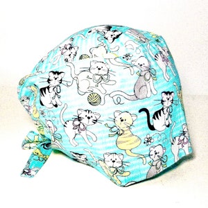 Może przedstawiać: Jasno niebieski czepek chirurgiczny z tkaniny z wzorem białych i szarych kotów z kokardkami. Koty znajdują się na jasnoniebieskim i białym paskowanym tle.