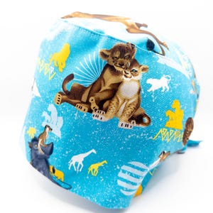 May include: A blue scrub hat with a Lion King print featuring Simba and Nala. The print also includes giraffes, zebras, and other animals from the film.