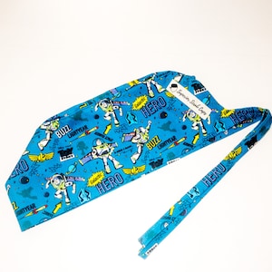 May include: A blue scrub cap featuring a repeating pattern of the character Buzz Lightyear from Toy Story, with the words "Buzz Lightyear" and "Hero" in a cartoon style. The cap has a matching tie and a tag that reads "Superior Scrub Caps."