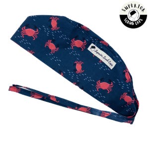 Peut inclure: Un bonnet de chirurgien bleu marine avec un motif répété de crabes rouges et de tirets blancs. Le bonnet a une attache à l'arrière et une petite étiquette blanche avec l'inscription "Superior Scrub Caps".