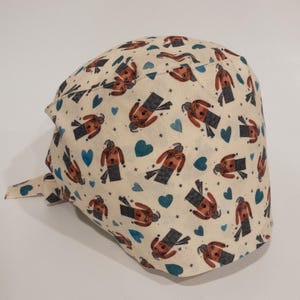 Könnte beinhalten: Eine cremefarbene Stoff-OP-Mütze mit einem sich wiederholenden Muster aus braunen Figuren mit Brille und blauen Herzen. Die Mütze hat einen seitlichen Knoten.