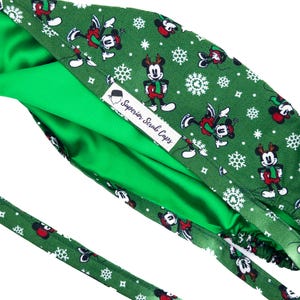 May include: Green scrub cap with a festive pattern featuring Mickey Mouse in holiday attire and snowflakes. The cap has a bright green interior and a label that reads "Superior Scrub Caps".