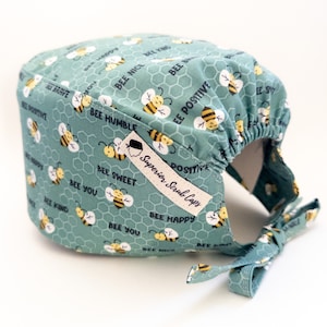 May include: A teal scrub cap with a repeating pattern of bees and honeycomb shapes. The cap features positive affirmations like "Bee Kind" and "Bee Happy." A white tag reads "Superior Scrub Caps."