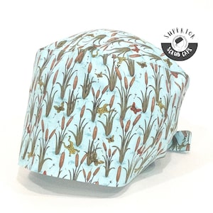 May include: A light blue scrub cap with a pattern of green reeds, brown cattails, and yellow frogs. The text "Superior Scrub Caps" is printed in black on a white circle.