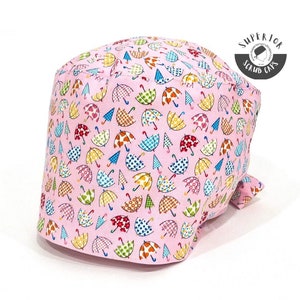Peut inclure: Un bonnet de travail rose avec un imprimé d'ombrelles colorées. Le bonnet a une attache à l'arrière et le texte "Superior Scrub Caps" est imprimé sur un cercle blanc.