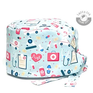 Könnte beinhalten: Eine hellblaue OP-Mütze mit einem Bindeband am Rücken. Die Mütze hat ein Muster aus medizinischen Symbolen und Icons, darunter ein Stethoskop, ein Herz, eine Erste-Hilfe-Box, eine Spritze und eine Pillenflasche. Der Text "Superior Scrub Caps" ist auf der Mütze aufgedruckt.