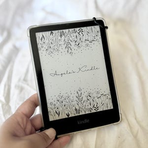 May include: A black Kindle e-reader with a clear protective case. The screen displays a floral design with the text "Angela's Kindle" and the brand name "Kindle".