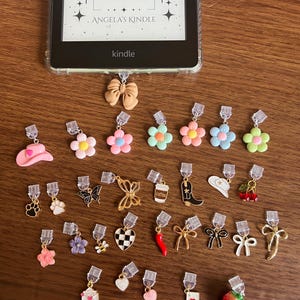 May include: A collection of decorative Kindle page holders in various shapes and colors, including flowers, bows, hearts, and a cowboy hat. The Kindle screen displays "Angela's Kindle" and the Kindle logo.