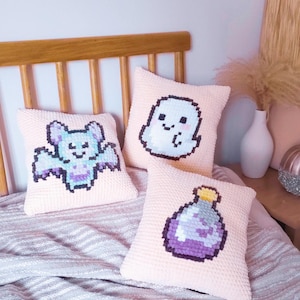 May include: Three pink square cushions with pixelated Halloween designs: a bat, a ghost, and a potion bottle. The text "KAWAII HALLOWEEN CUSHION" and "SIX ADORABLE CROCHET & CROSS-STITCH PATTERNS" are visible. Designed by I Dilly Dally.