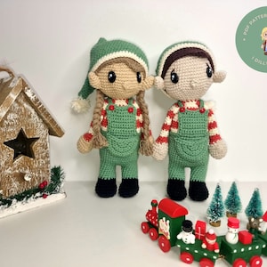 Op de afbeelding: Twee gehaakte elfpoppen met groene overalls, rood-wit gestreepte shirts en bruin haar. De poppen staan op een wit oppervlak met een houten vogelhuisje en een speelgoedtreinset.
