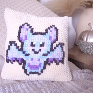May include: A square, cream-coloured cushion featuring a pixelated bat design in shades of purple, blue, and grey. The bat has outstretched wings and a smiling face. The cushion has a textured, plush surface.