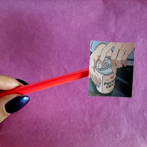 Może przedstawiać: Zbliżenie na rękę otwierającą puszkę napoju "PREMIUM CHAI". Puszka jest srebrna z czerwoną i białą etykietą. Ręka jest narysowana w stylu anime.