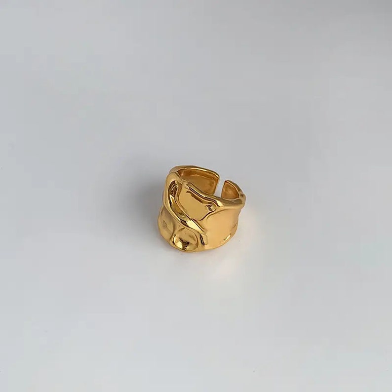 K&ouml;nnte beinhalten: Ein goldfarbener Ring mit einem strukturierten, abstrakten Design. Der Ring ist offen und verstellbar.