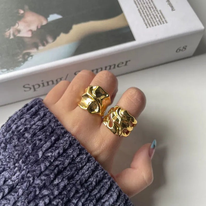 K&ouml;nnte beinhalten: Zwei goldfarbene Ringe mit abstraktem, organischem Design. Die Ringe werden am Zeige- und Mittelfinger einer Hand getragen.