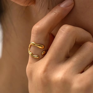 May include: A gold-colored ring with an organic, flowing design. The ring features a curved shape with rounded, droplet-like elements. The ring is worn on a finger, showcasing its unique, modern aesthetic. The jewelry piece has a polished, reflective surface.