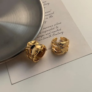 K&ouml;nnte beinhalten: Zwei goldfarbene Ringe mit abstraktem, strukturiertem Design. Die Ringe sind aus Metall und haben einen klobigen, modernen Look.